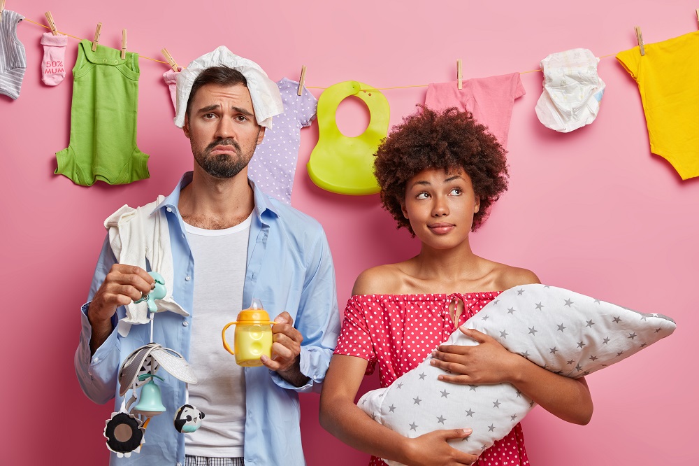 sad tired father poses near thoughtful wife with baby on hands take care of newborn being tired of parenthood pose against pink background rope with washed childs items. young family concept